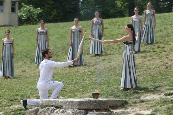 扮演最高女祭司的演員瑪麗·米娜（前右）點燃第一棒火炬手——希臘運動員、東京奧運會男子賽艇項目金牌得主斯特凡諾斯·恩圖斯科斯手中的火炬。新華社記者 趙丁喆 攝